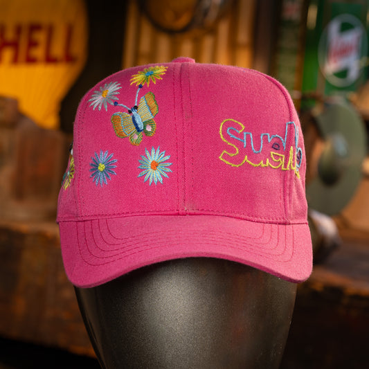 Pink cap with floral designs and 'SueMe' text on a blurred background