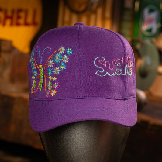 Purple cap with butterfly and floral design and embroidered name on a blurred background