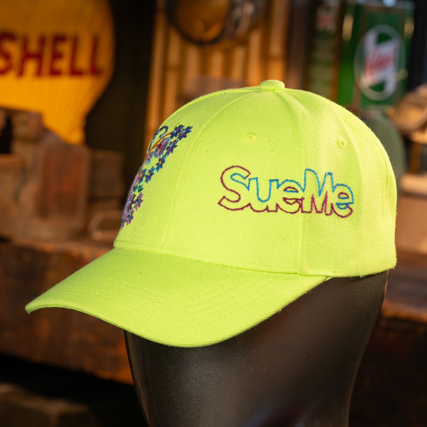 Yellow cap with colorful 'SueMe' embroidery on a mannequin head against a blurred background.
