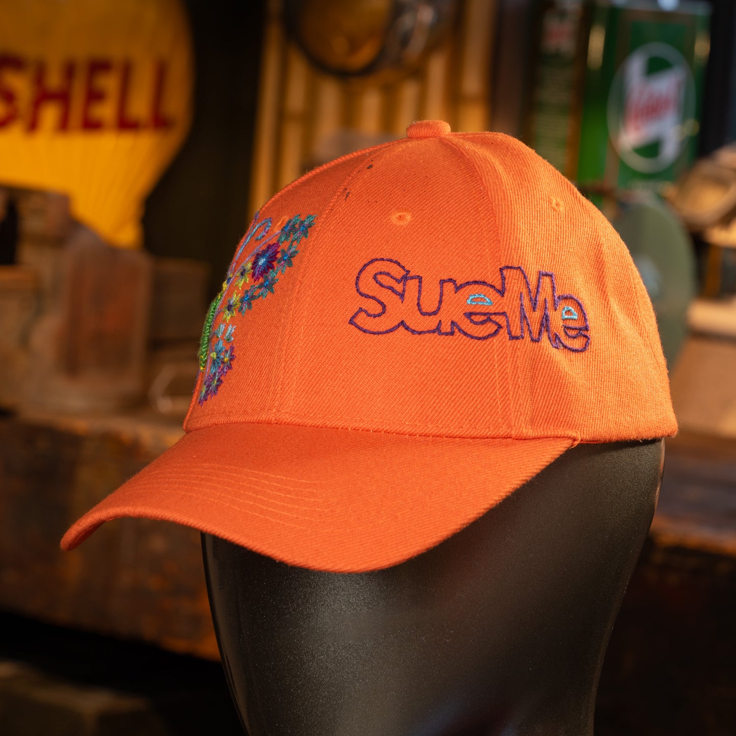 Orange cap with 'Sue Me' embroidery on a mannequin head, blurred background