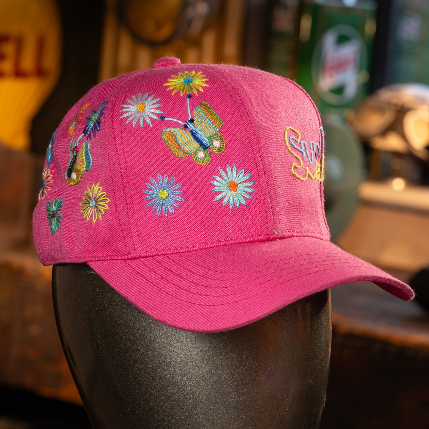 Pink cap with colorful butterfly and flower designs on a mannequin head against a blurred background.