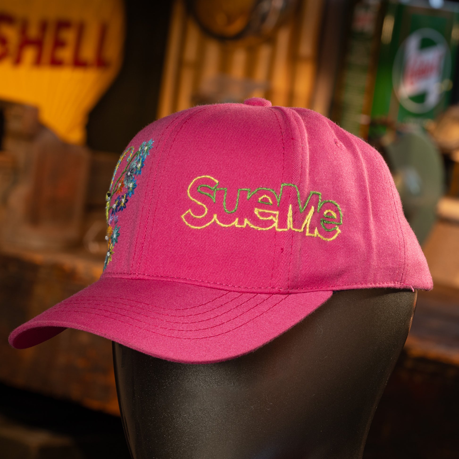Pink cap with 'SueMe' embroidery on a mannequin head, blurred background