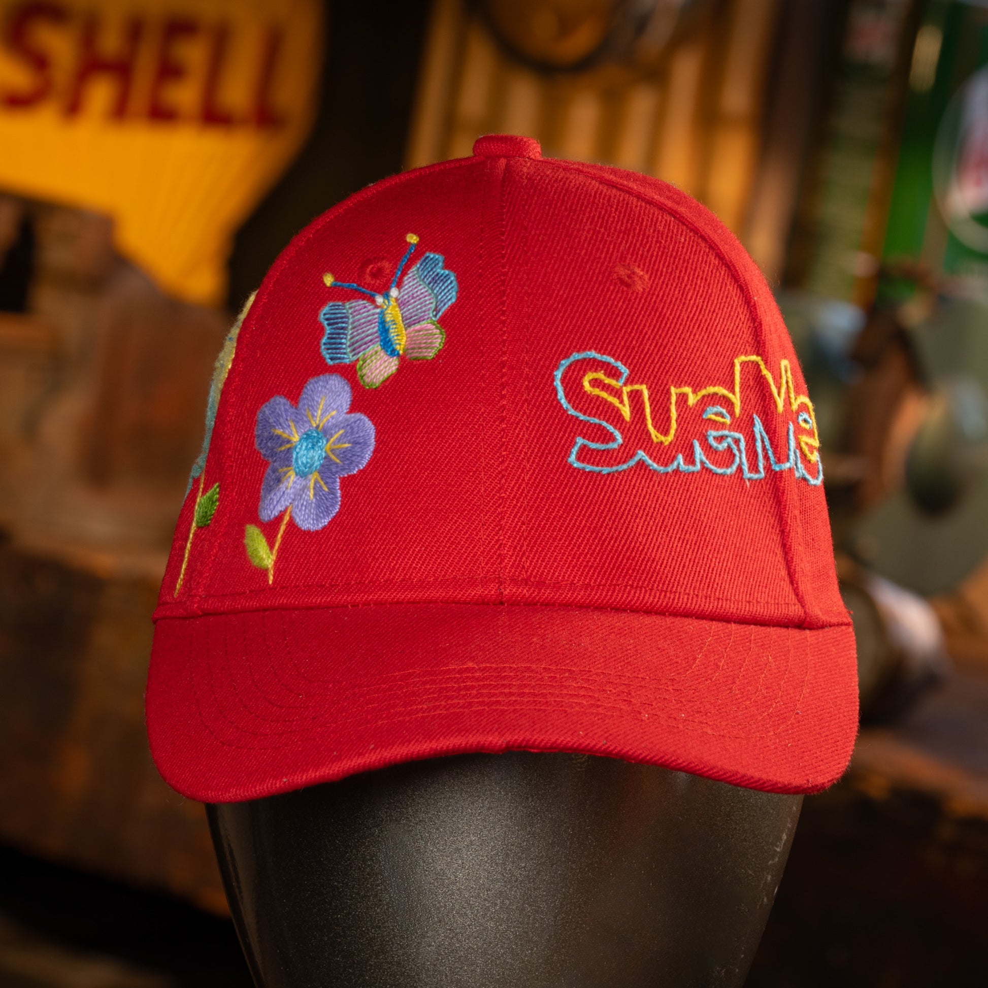 Red cap with embroidered flowers and 'SueMe' text on a blurred background
