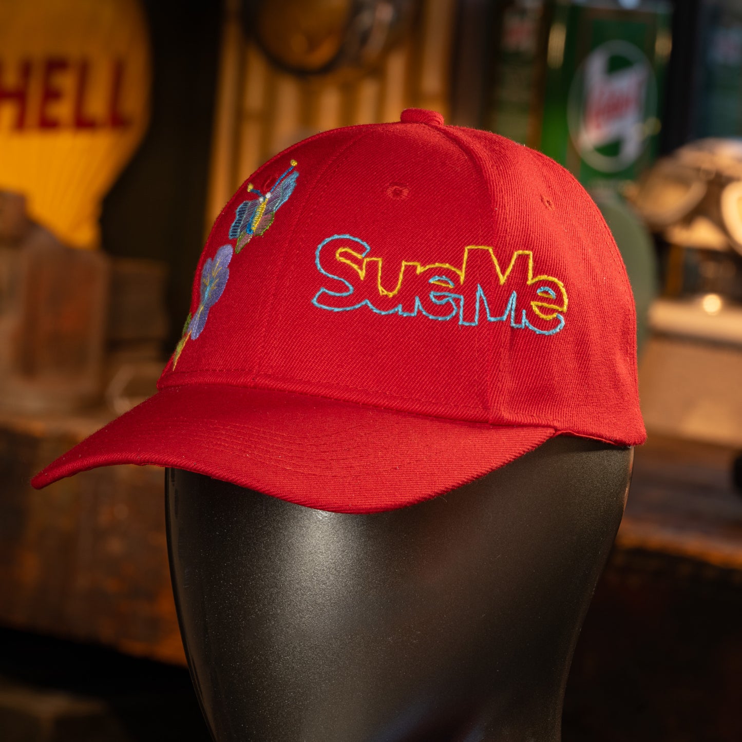 Red cap with 'SueMe' embroidery on a mannequin head in a bar setting.