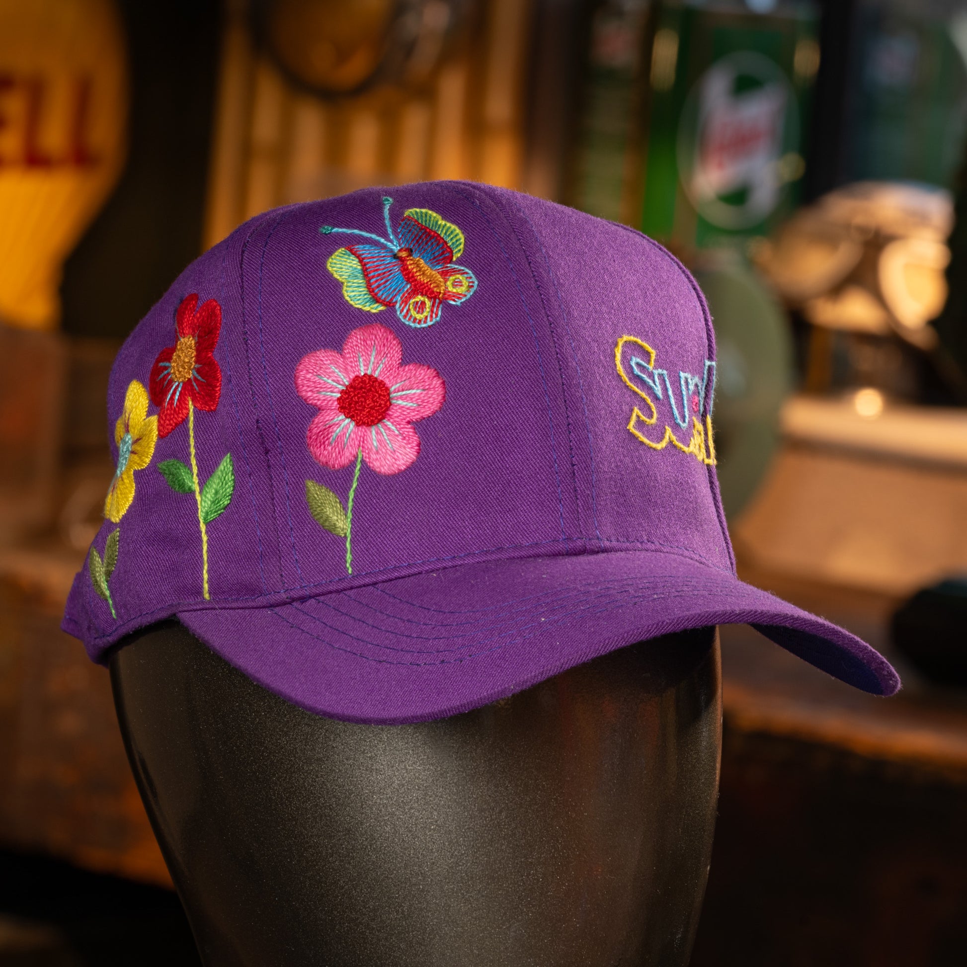 Purple cap with floral and butterfly embroidery on a mannequin head against a blurred background.