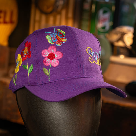 Purple cap with floral and butterfly embroidery on a mannequin head against a blurred background.