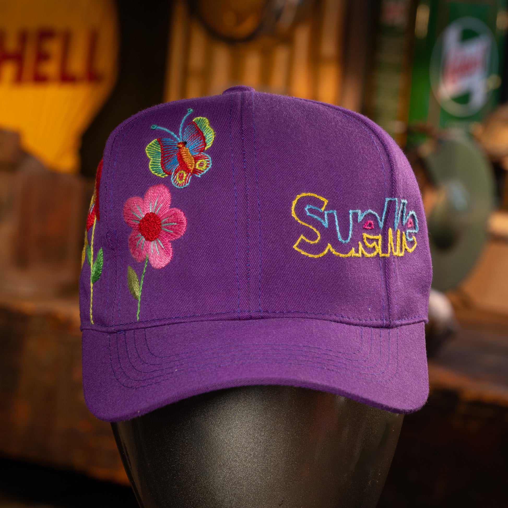 Purple cap with floral and butterfly embroidery on a mannequin head, with a blurred background.