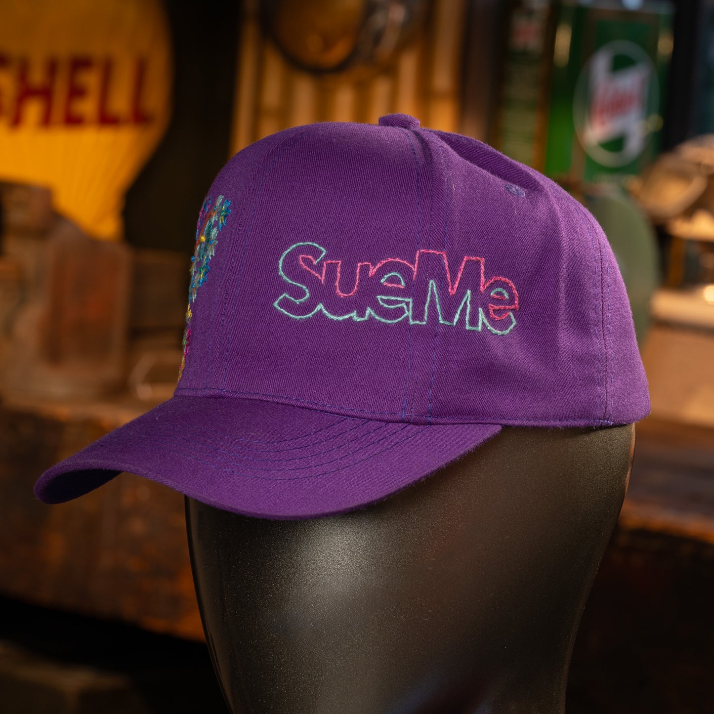 Purple cap with 'Sue Me' embroidery on a mannequin head in a bar setting.