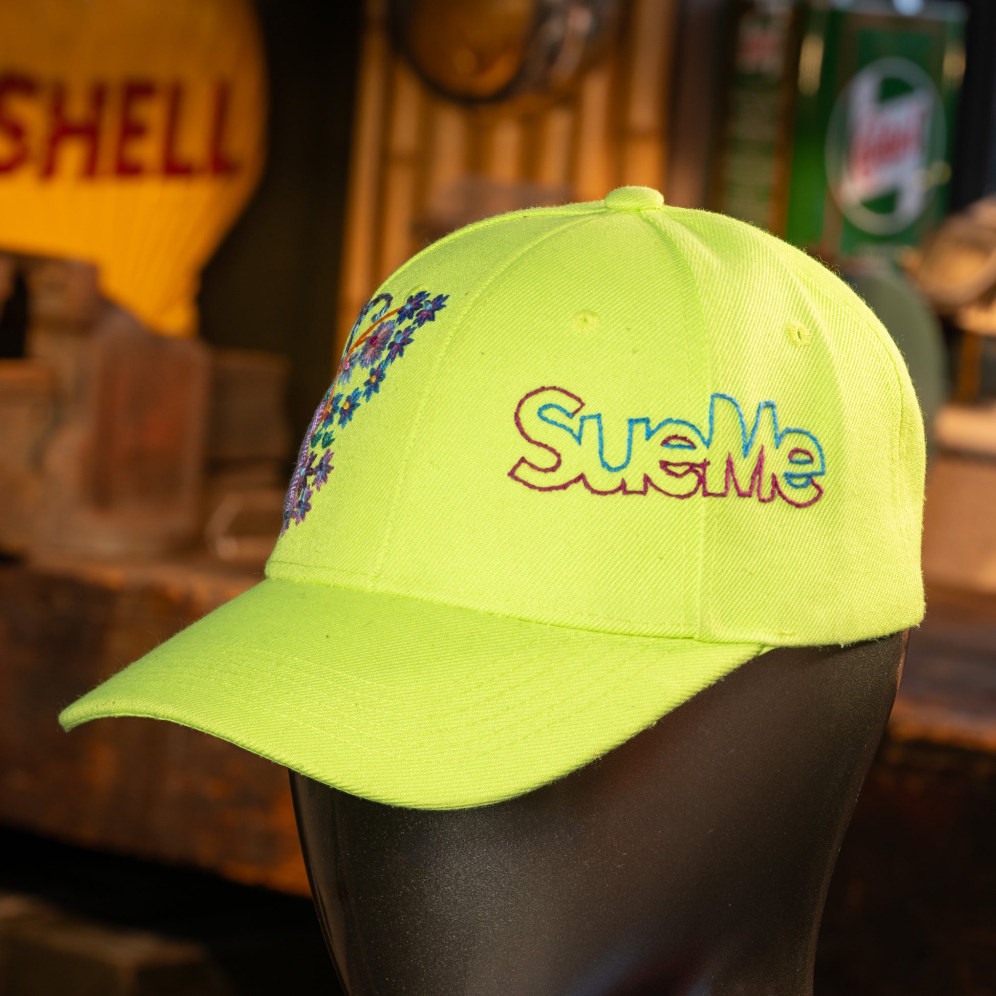 Yellow cap with colorful 'SueMe' embroidery on a mannequin head against a blurred background.