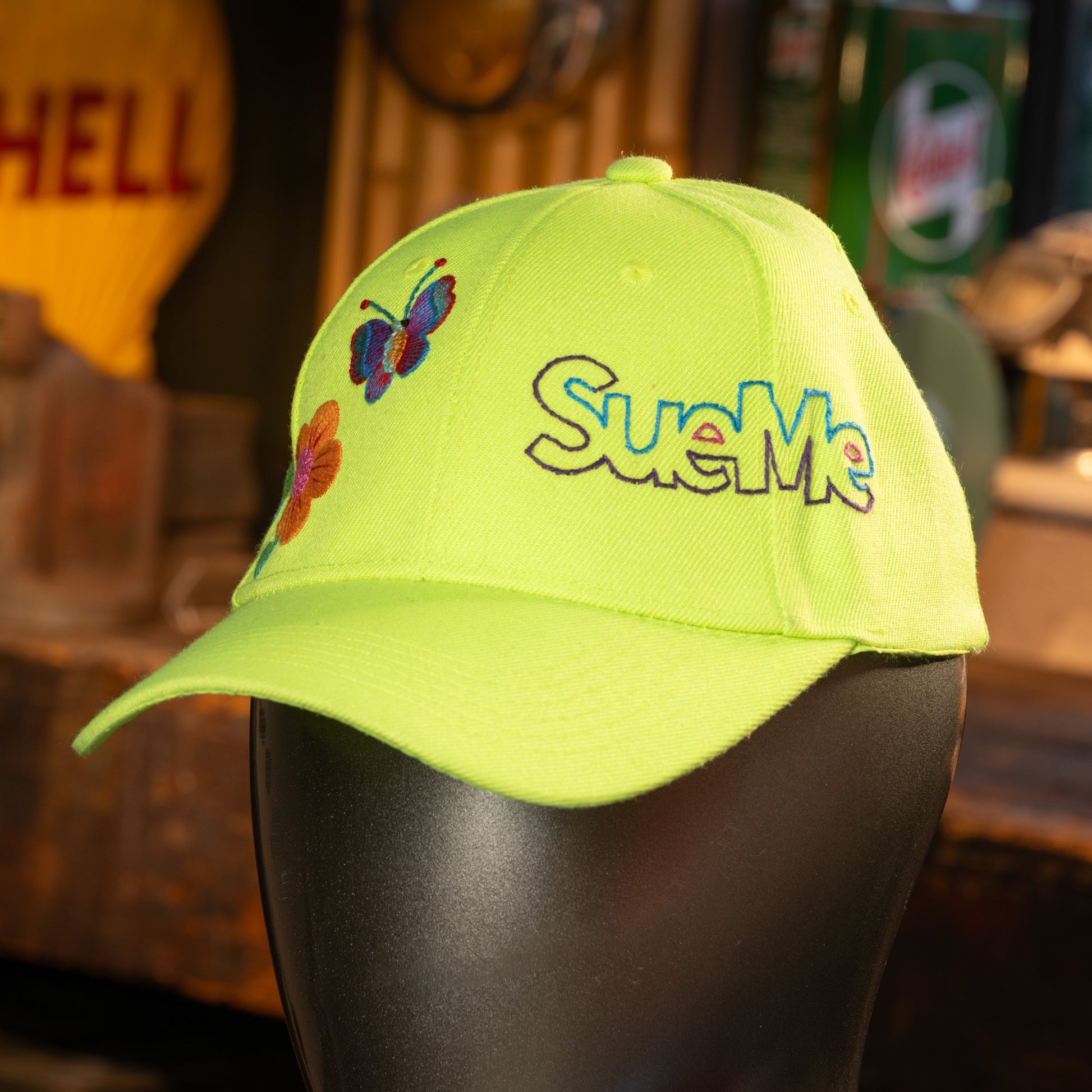 Neon green cap with 'SueMe' text and butterfly design on a mannequin head.