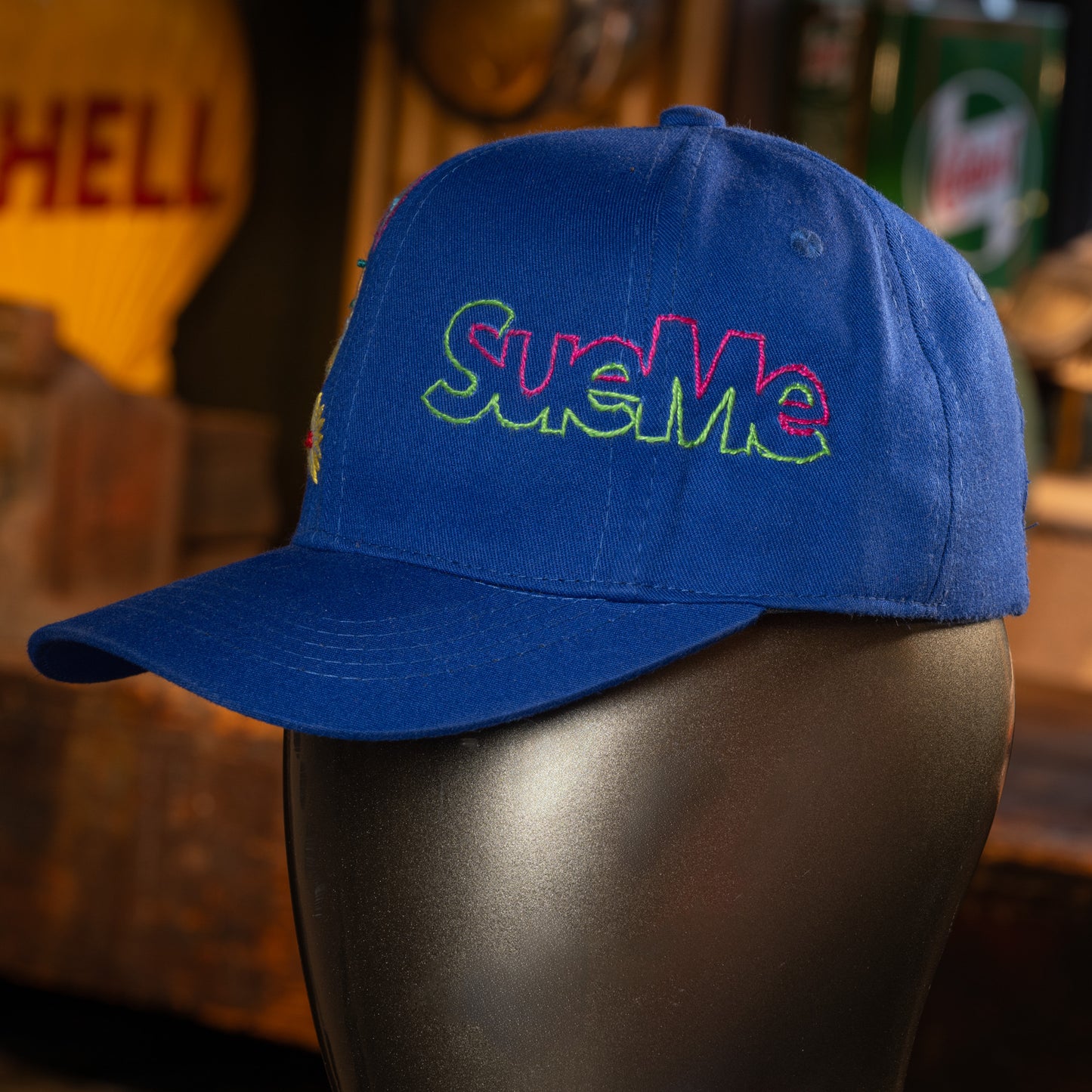 Blue cap with colorful 'Sue Me' text on a mannequin head against a blurred background