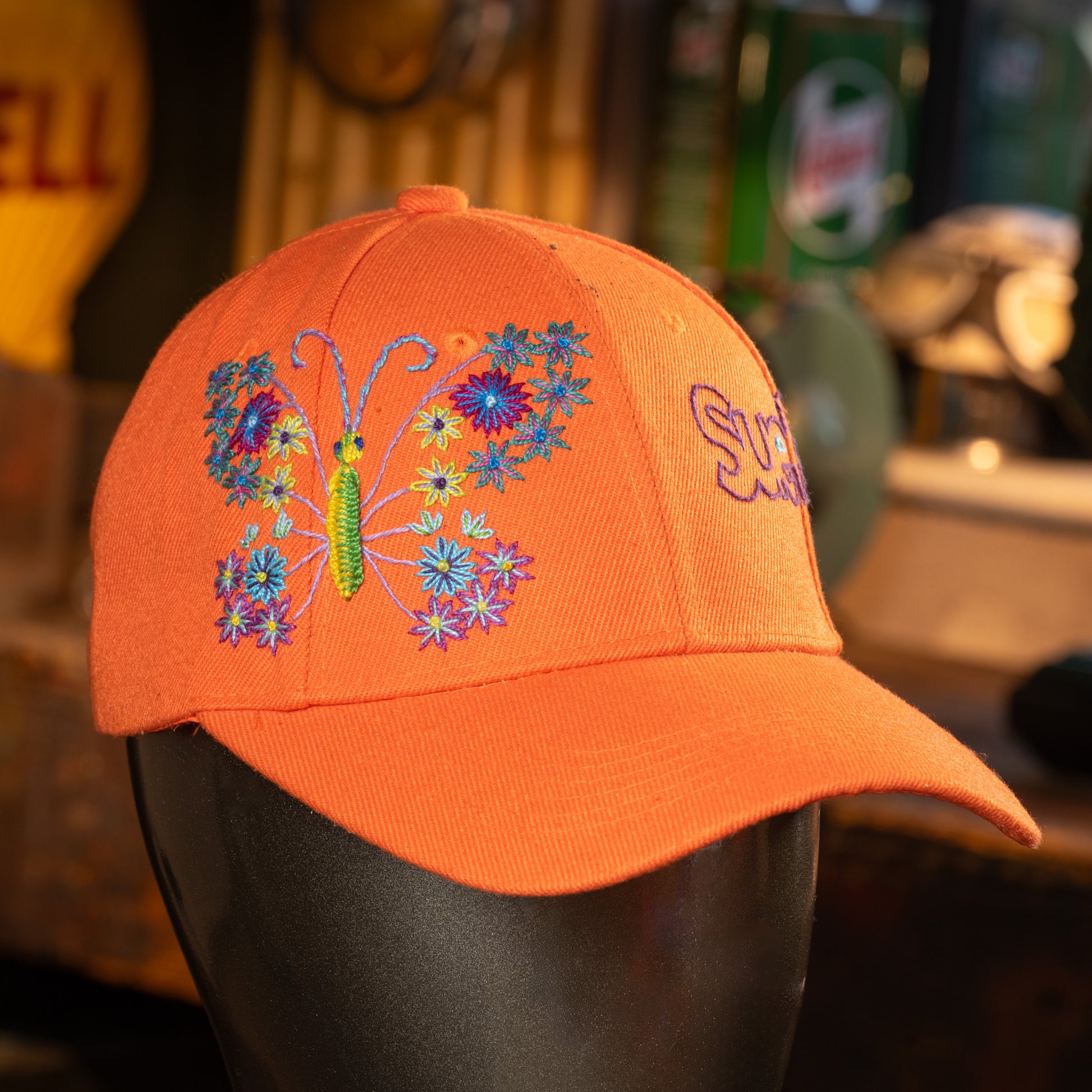 Orange cap with butterfly and floral design on a mannequin head against a blurred background.