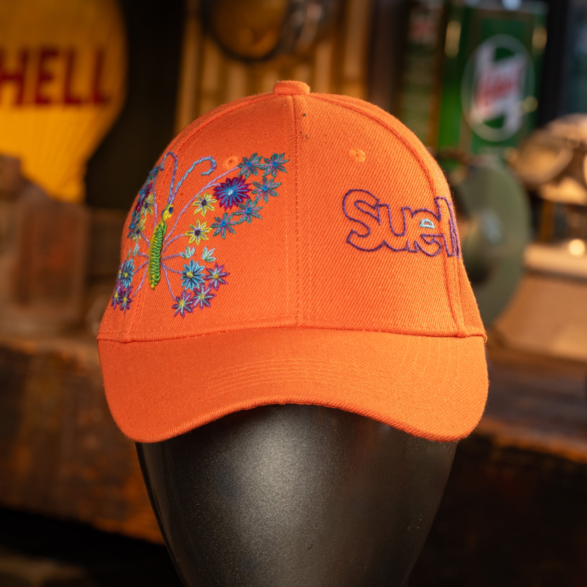Orange cap with floral embroidered design and 'SueMe' text on a mannequin head against a blurred background.