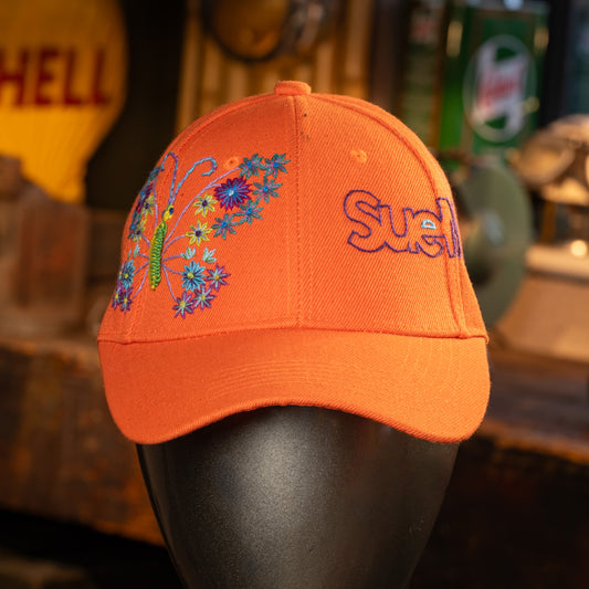 Orange cap with floral embroidered design and 'SueMe' text on a mannequin head against a blurred background.